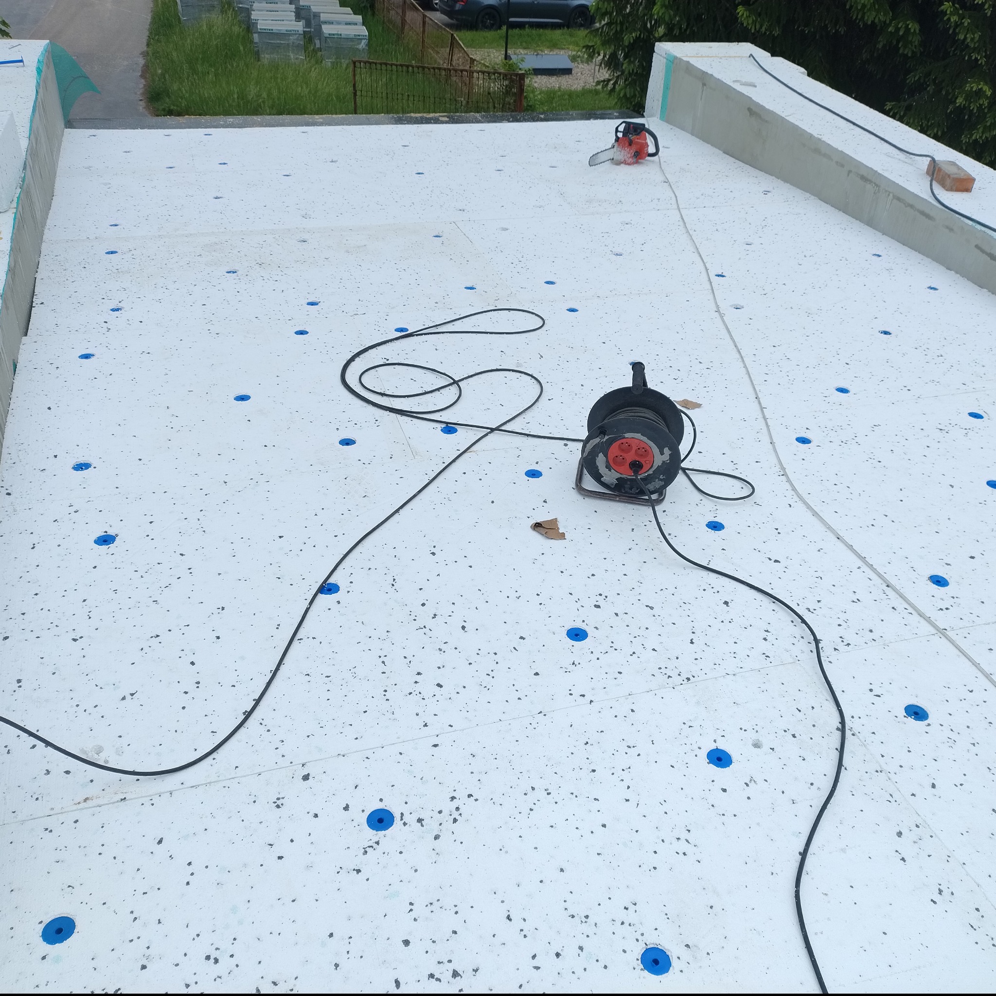 Ocieplenie dachu z pianki z widocznymi niebieskimi kołkami mocującymi, przedłużaczem i piłą łańcuchową. Widok z góry na płaski dach.