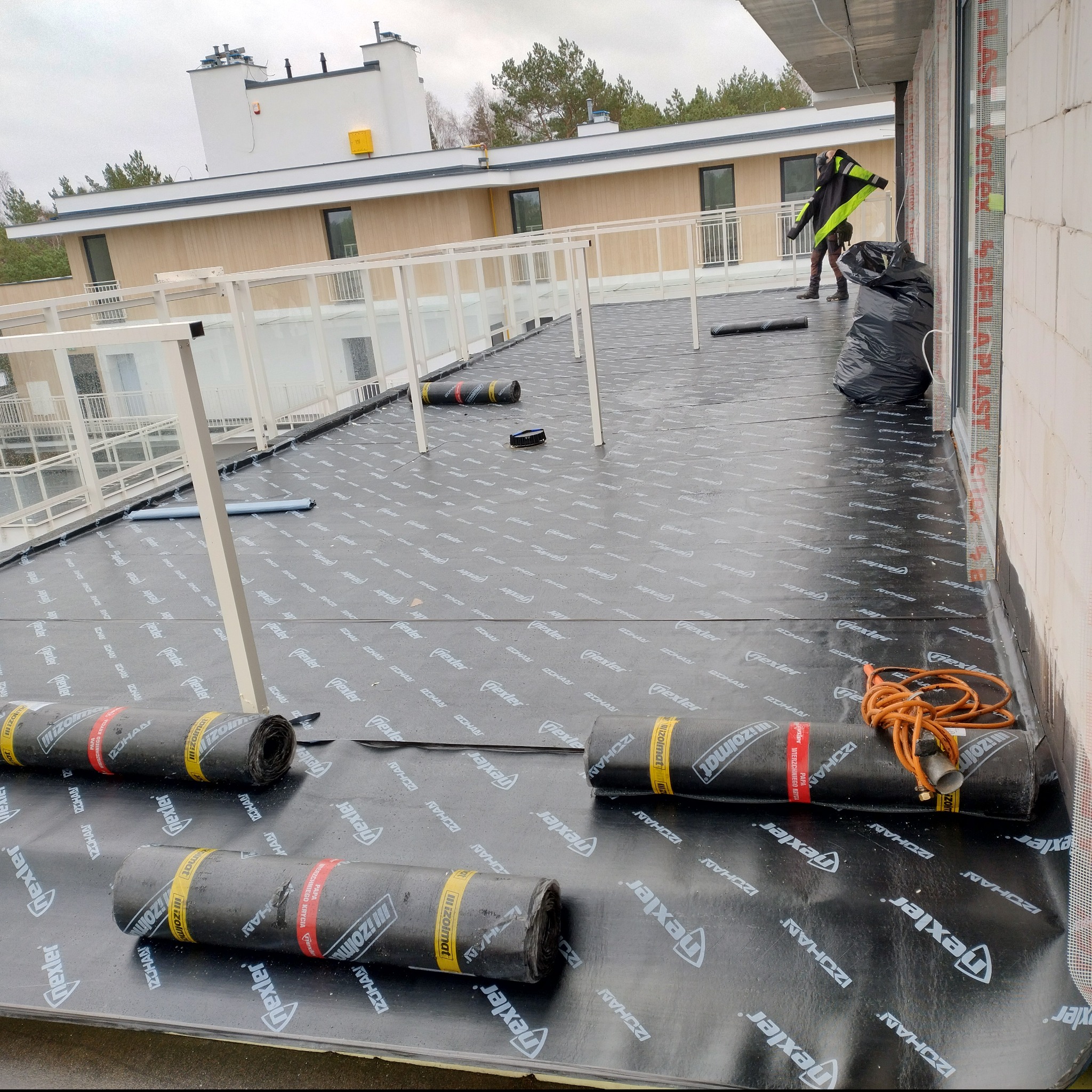 Prace dekarskie: papa termozgrzewalna na dachu budynku wielorodzinnego. Widoczny pracownik w odblaskowej kurtce i zgrzewarka.