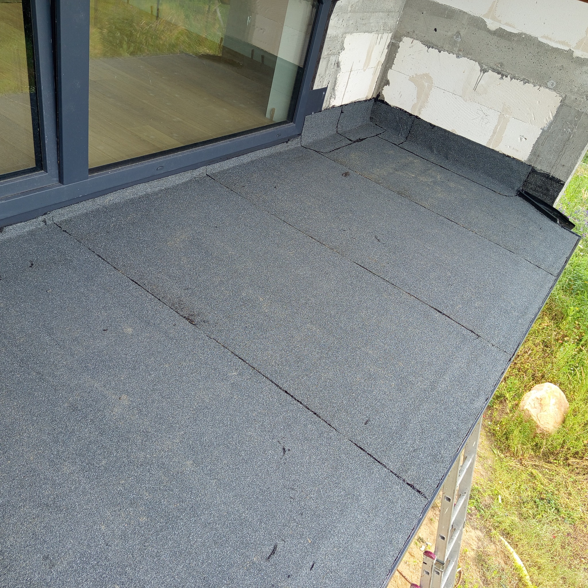 Szary dach płaski pokryty papą termozgrzewalną, widoczny fragment okna dachowego, ściana z betonu komórkowego i fragment drabiny w tle.