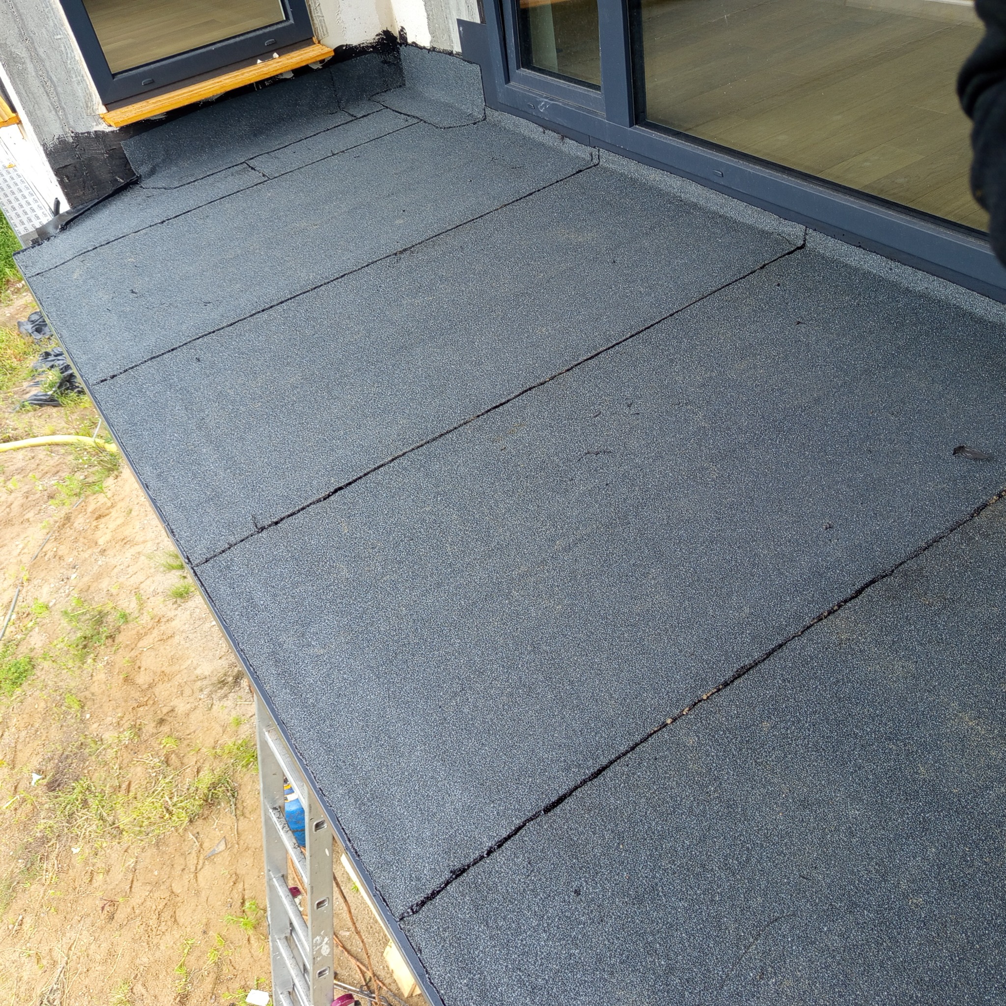 Szczegółowe ujęcie dachu pokrytego papą termozgrzewalną, z widocznymi łączeniami i oknem dachowym. Układ połaci w regularnych rzędach.