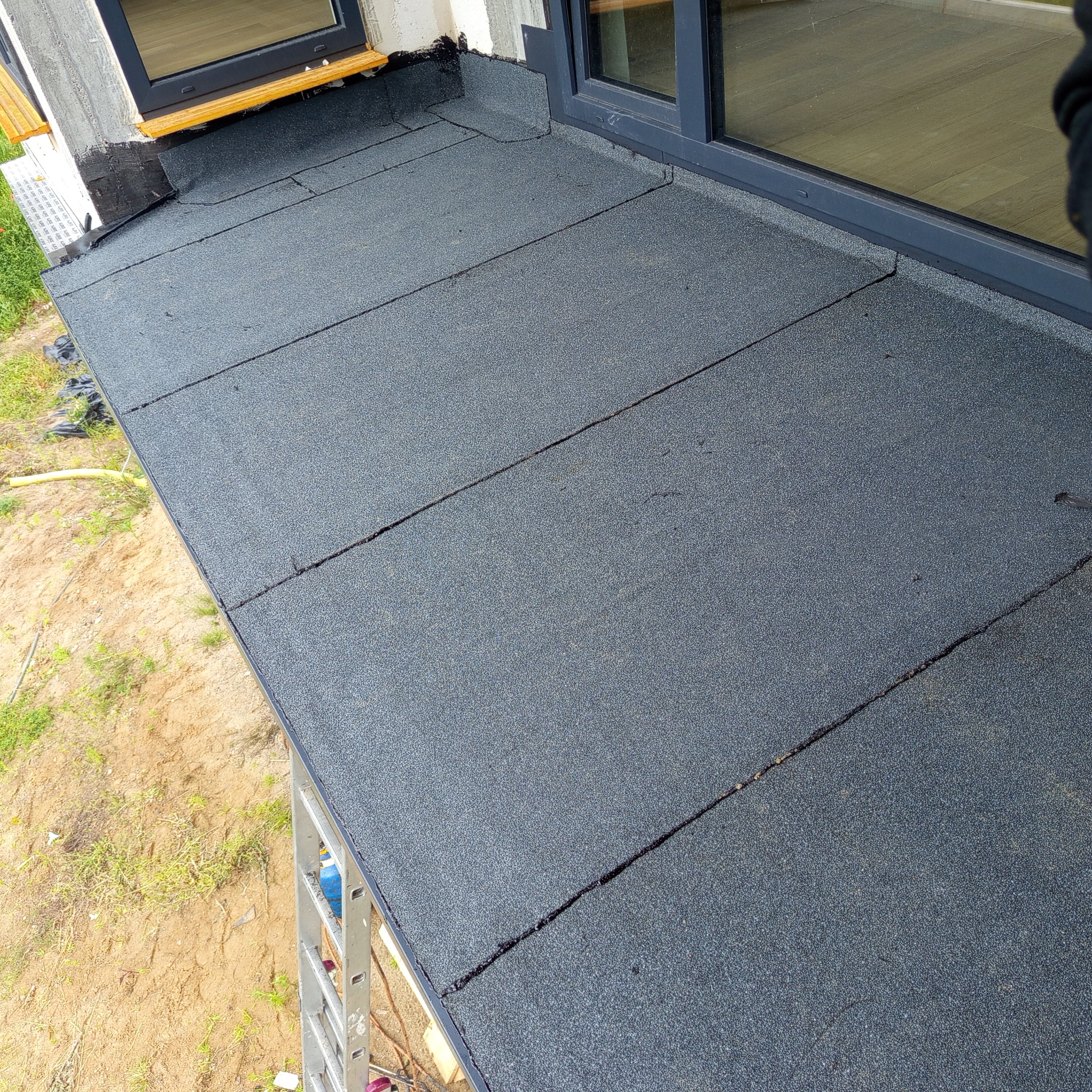 Wykończony dach płaski z papy bitumicznej przy oknie, widok z góry. Układ papy z widocznymi łączeniami. Fragment drabiny w rogu kadru.