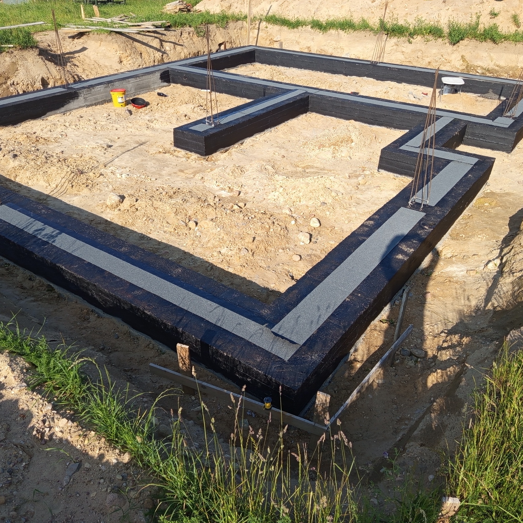 Izolacja fundamentów z papy i styroduru na piaskowym podłożu, z widocznymi prętami zbrojeniowymi i żółtym pojemnikiem. Ujęcie z góry, słoneczny dzień.