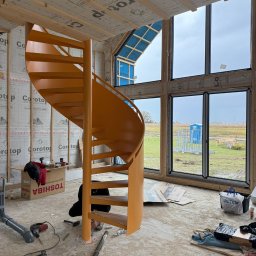 STALUX SPÓŁKA Z OGRANICZONĄ ODPOWIEDZIALNOŚCIĄ - Montaż pomarańczowych schodów spiralnych w surowym wnętrzu domu w trakcie budowy. Widoczne narzędzia i materiały budowlane. Duże okna z widokiem na zewnątrz.