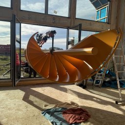 STALUX SPÓŁKA Z OGRANICZONĄ ODPOWIEDZIALNOŚCIĄ - Montaż spiralnych schodów w surowym wnętrzu. Żółte schody podnoszone przez maszynę, widoczne przez otwarte okno na tle krajobrazu. Na podłodze leżą szmaty.