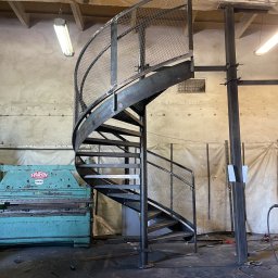 STALUX SPÓŁKA Z OGRANICZONĄ ODPOWIEDZIALNOŚCIĄ - Metalowe schody spiralne z siatkową balustradą w warsztacie w Toszku. Surowy metal, widoczne ślady obróbki. W tle maszyna do obróbki metalu i elementy konstrukcyjne.