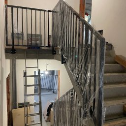 STALUX SPÓŁKA Z OGRANICZONĄ ODPOWIEDZIALNOŚCIĄ - Czarne, metalowe schody z balustradą w folii ochronnej, w surowym wnętrzu. Widoczna drabina i elementy instalacji. Ujęcie z dołu, podkreślające konstrukcję schodów.