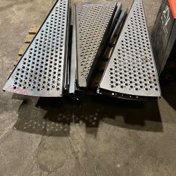 STALUX SPÓŁKA Z OGRANICZONĄ ODPOWIEDZIALNOŚCIĄ - Metalowe stopnie schodowe z perforacją, ułożone na palecie w warsztacie. Widoczne detale wykończenia i otwory antypoślizgowe. Surowy, industrialny charakter.