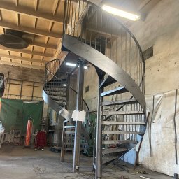 STALUX SPÓŁKA Z OGRANICZONĄ ODPOWIEDZIALNOŚCIĄ - Metalowe schody spiralne w warsztacie, surowy metal, widoczne spawy, oświetlenie warsztatowe, narzędzia w tle, wysoki kąt ujęcia, ukazujący konstrukcję w trakcie montażu.