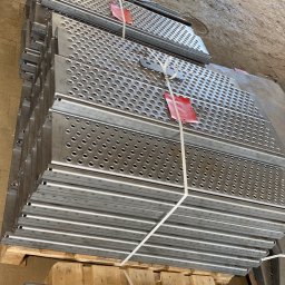 STALUX SPÓŁKA Z OGRANICZONĄ ODPOWIEDZIALNOŚCIĄ - Stos metalowych, perforowanych stopni na palecie, zabezpieczonych sznurkiem i oznaczonych czerwoną etykietą. Widoczne otwory i krawędzie, surowy wygląd.