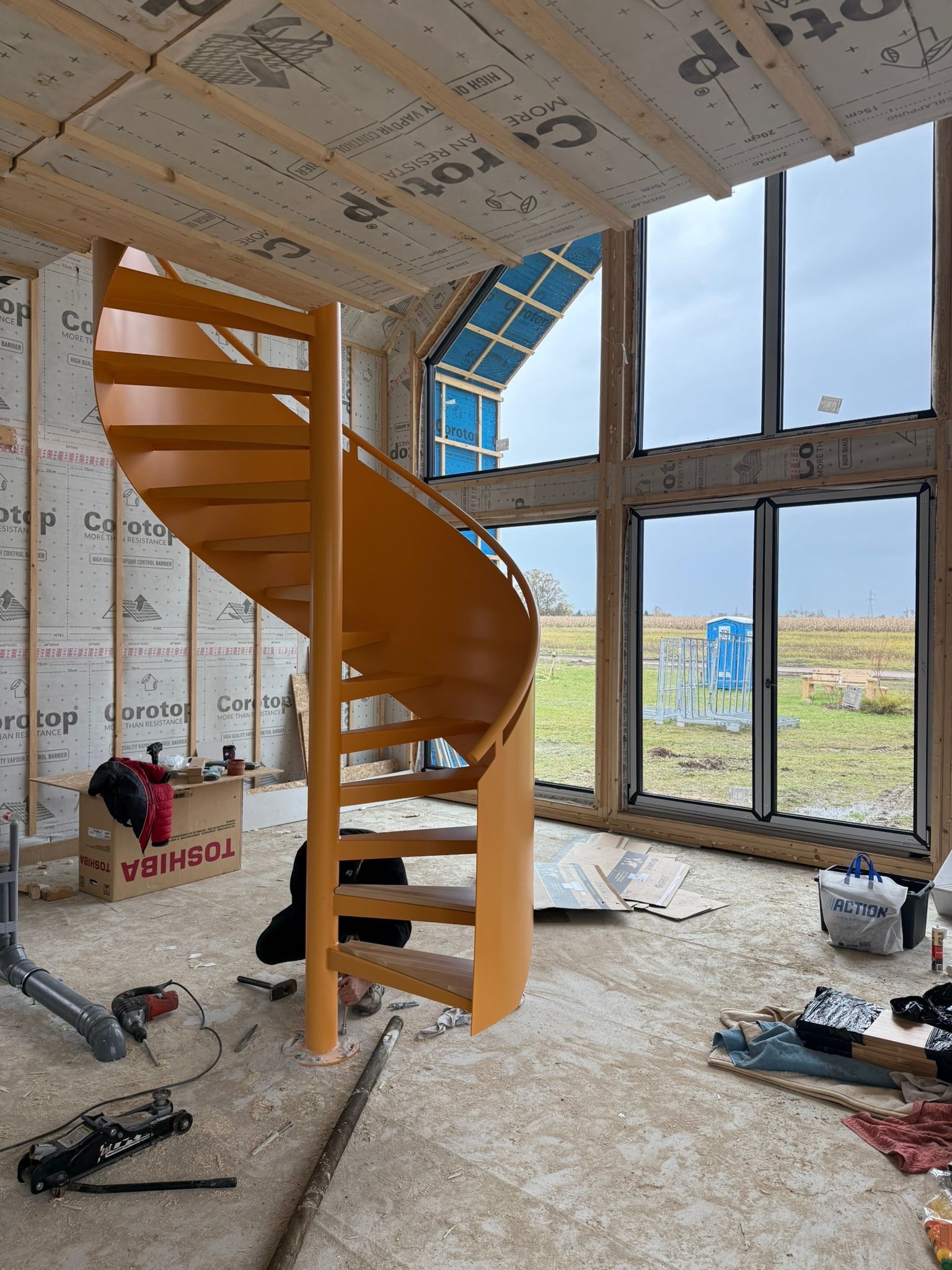 Montaż pomarańczowych schodów spiralnych w surowym wnętrzu domu w trakcie budowy. Widoczne narzędzia i materiały budowlane. Duże okna z widokiem na zewnątrz.