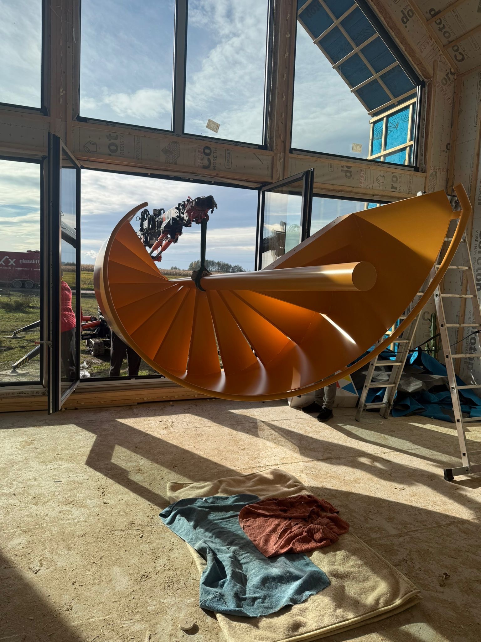 Montaż spiralnych schodów w surowym wnętrzu. Żółte schody podnoszone przez maszynę, widoczne przez otwarte okno na tle krajobrazu. Na podłodze leżą szmaty.