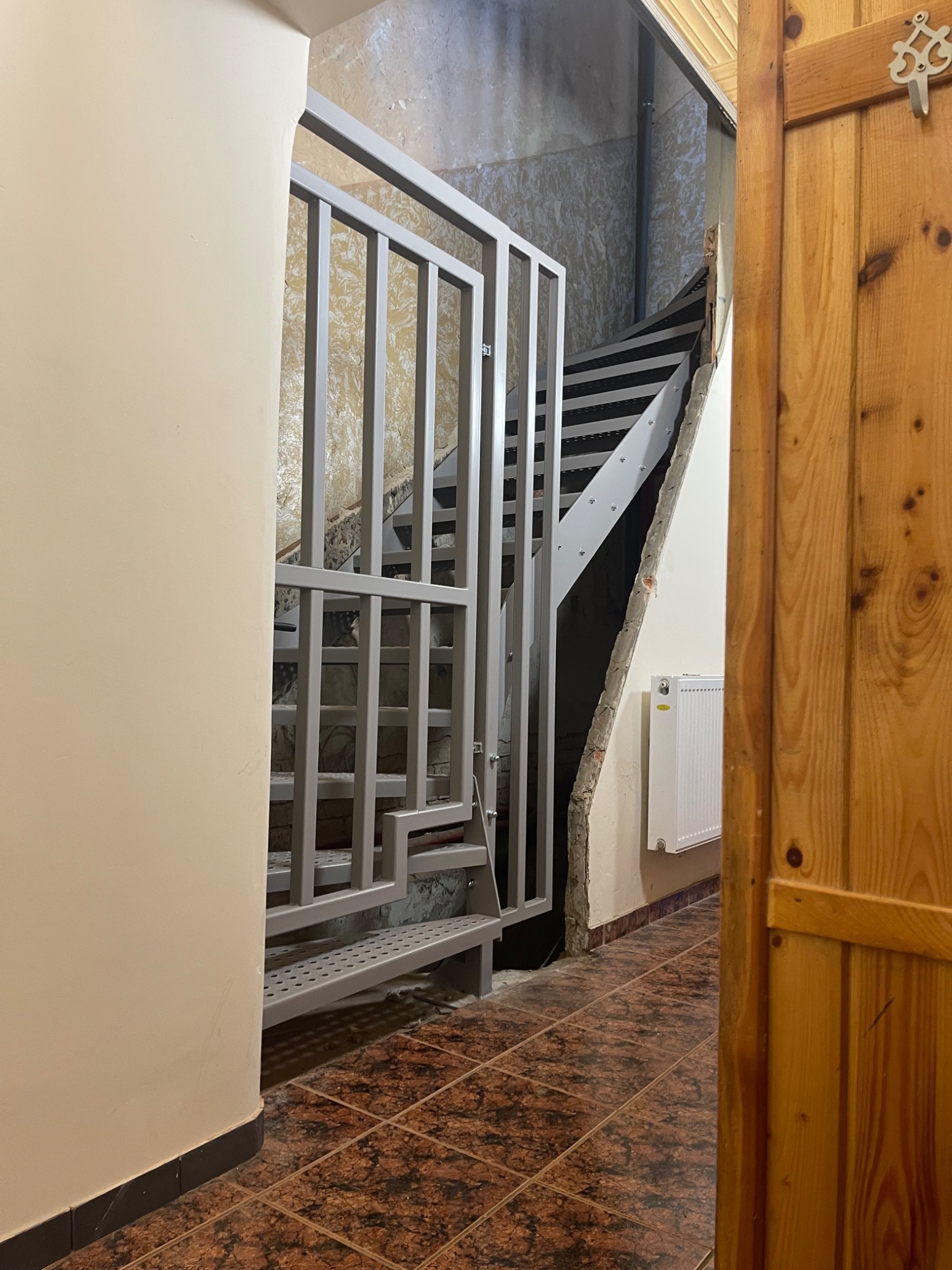 Metalowe schody z szarą, ażurową balustradą wewnątrz budynku, widoczne zza drewnianych drzwi. Brązowa podłoga z płytek. Ściany w odcieniach beżu i szarości.
