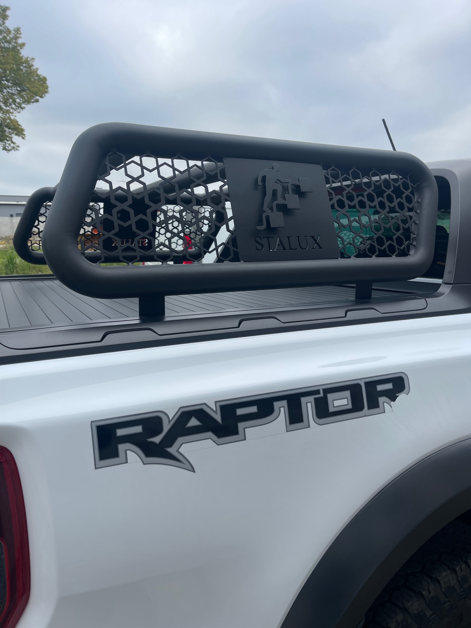 Czarna, ażurowa konstrukcja z logo STALUX zamontowana na pace białego pickupa Ford Raptor. Widoczny fragment tylnej lampy i napis RAPTOR.
