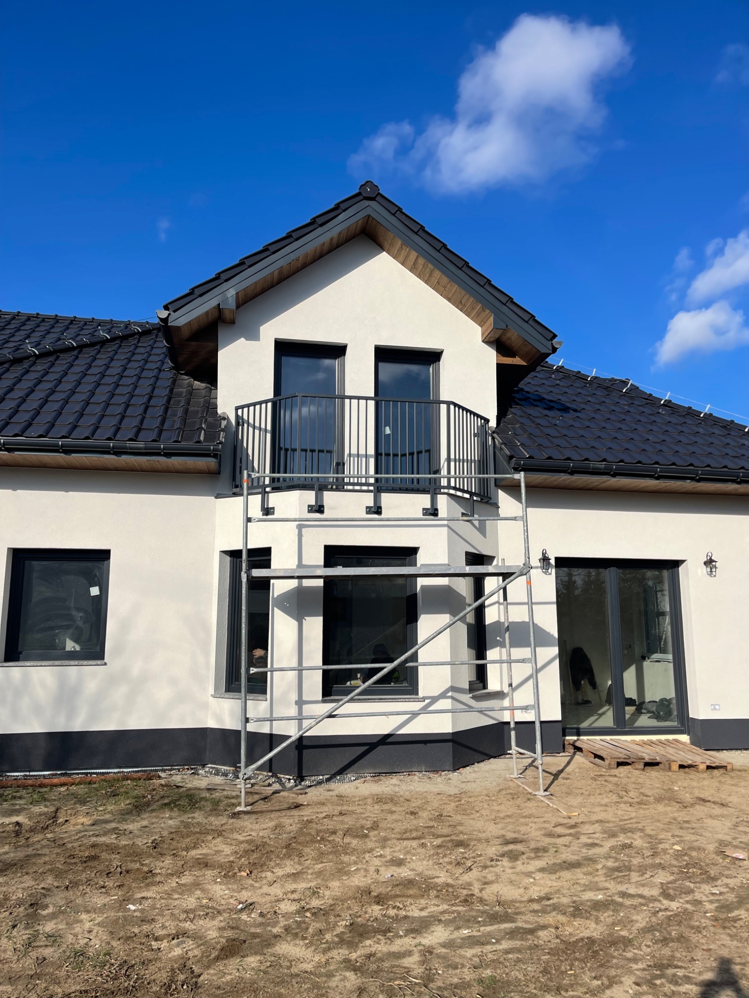 Nowoczesny dom z balkonem i czarną balustradą, widoczny ze stojącego rusztowania. Biała elewacja, ciemna dachówka i błękitne niebo w tle.