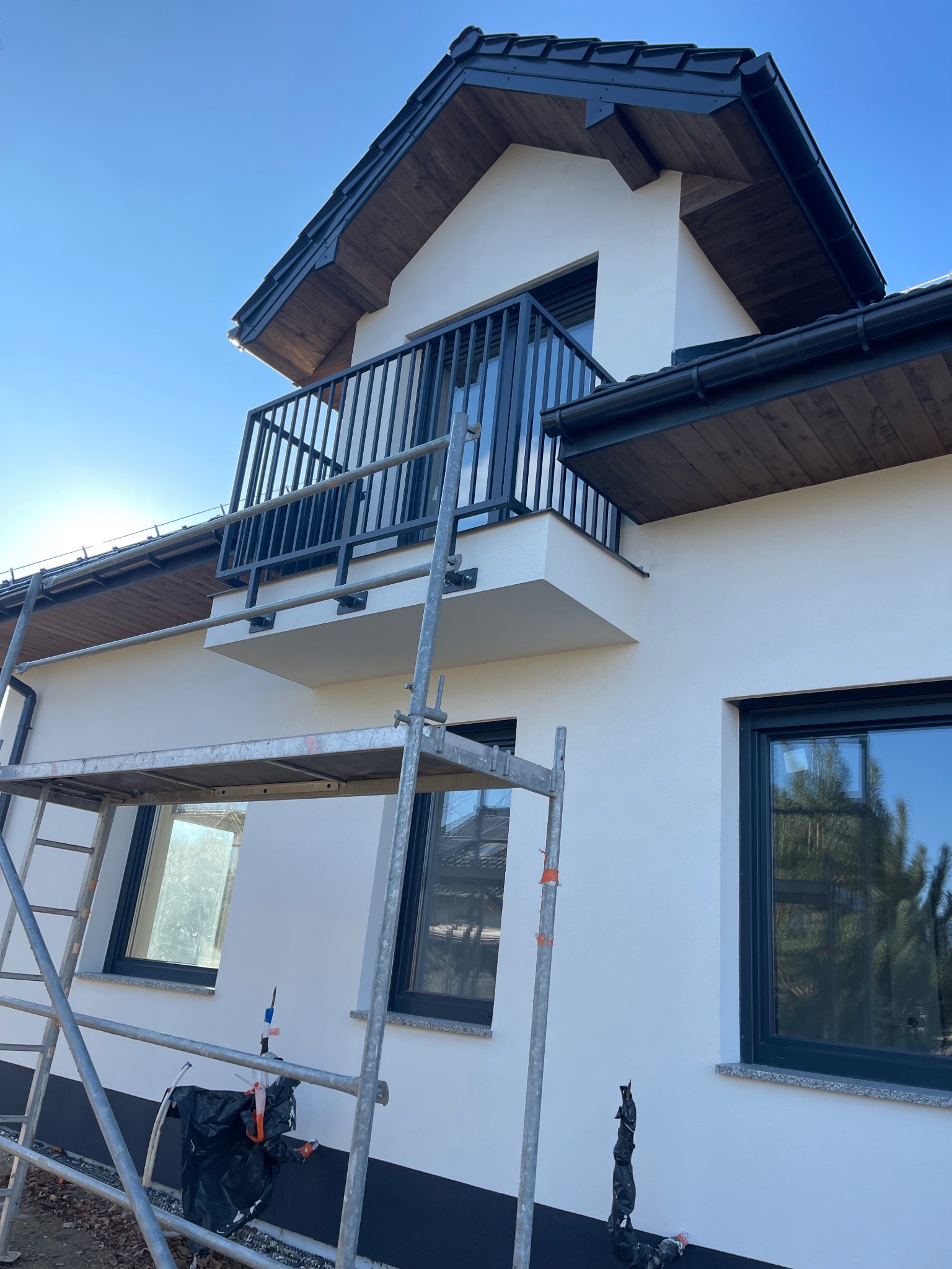 Nowoczesny balkon z czarną, metalową balustradą w nowym domu. Widoczne rusztowanie sugeruje prace wykończeniowe. Jasne tło nieba i drewniane elementy dachu.