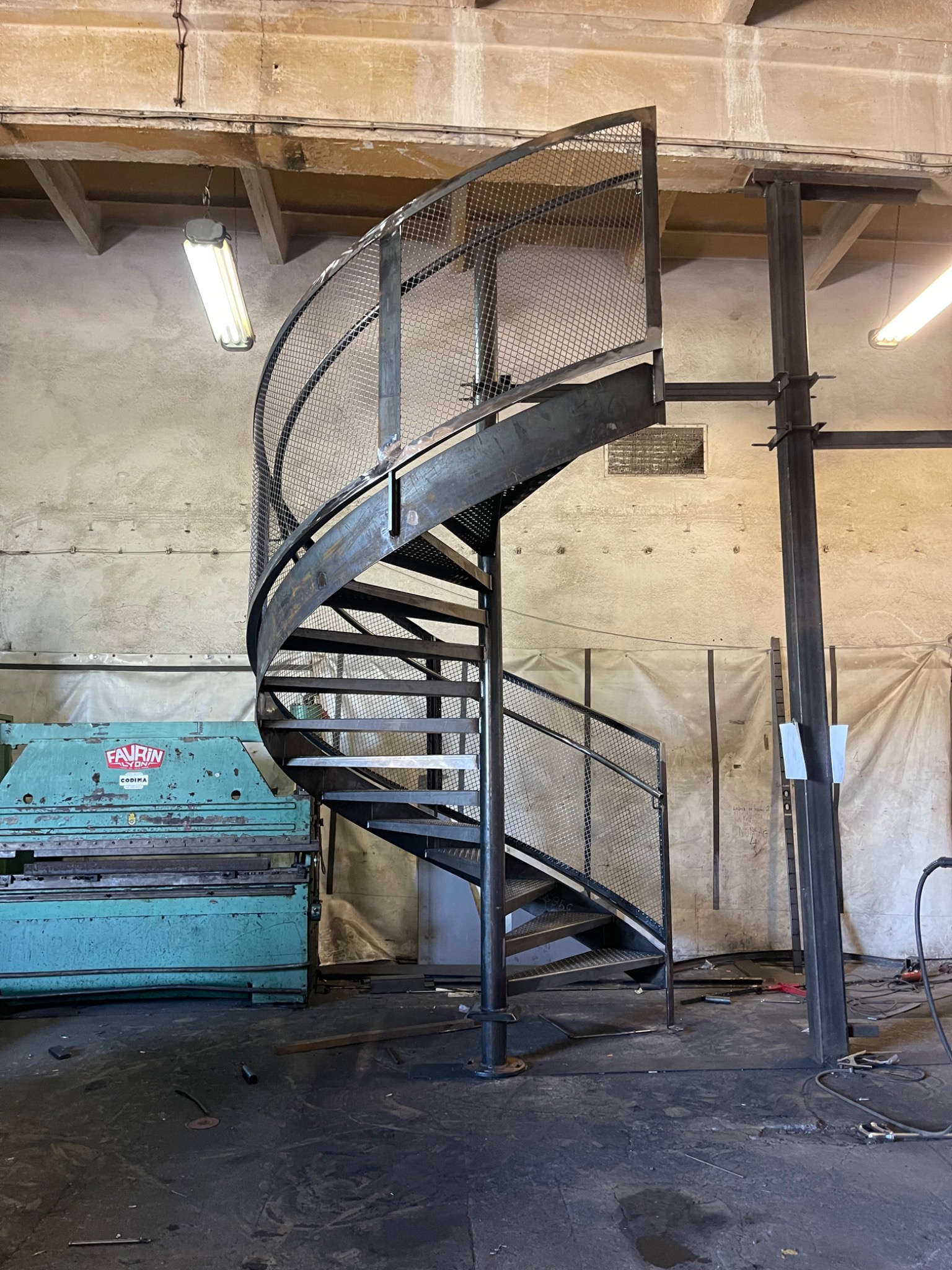 Metalowe schody spiralne z siatkową balustradą w warsztacie w Toszku. Surowy metal, widoczne ślady obróbki. W tle maszyna do obróbki metalu i elementy konstrukcyjne.