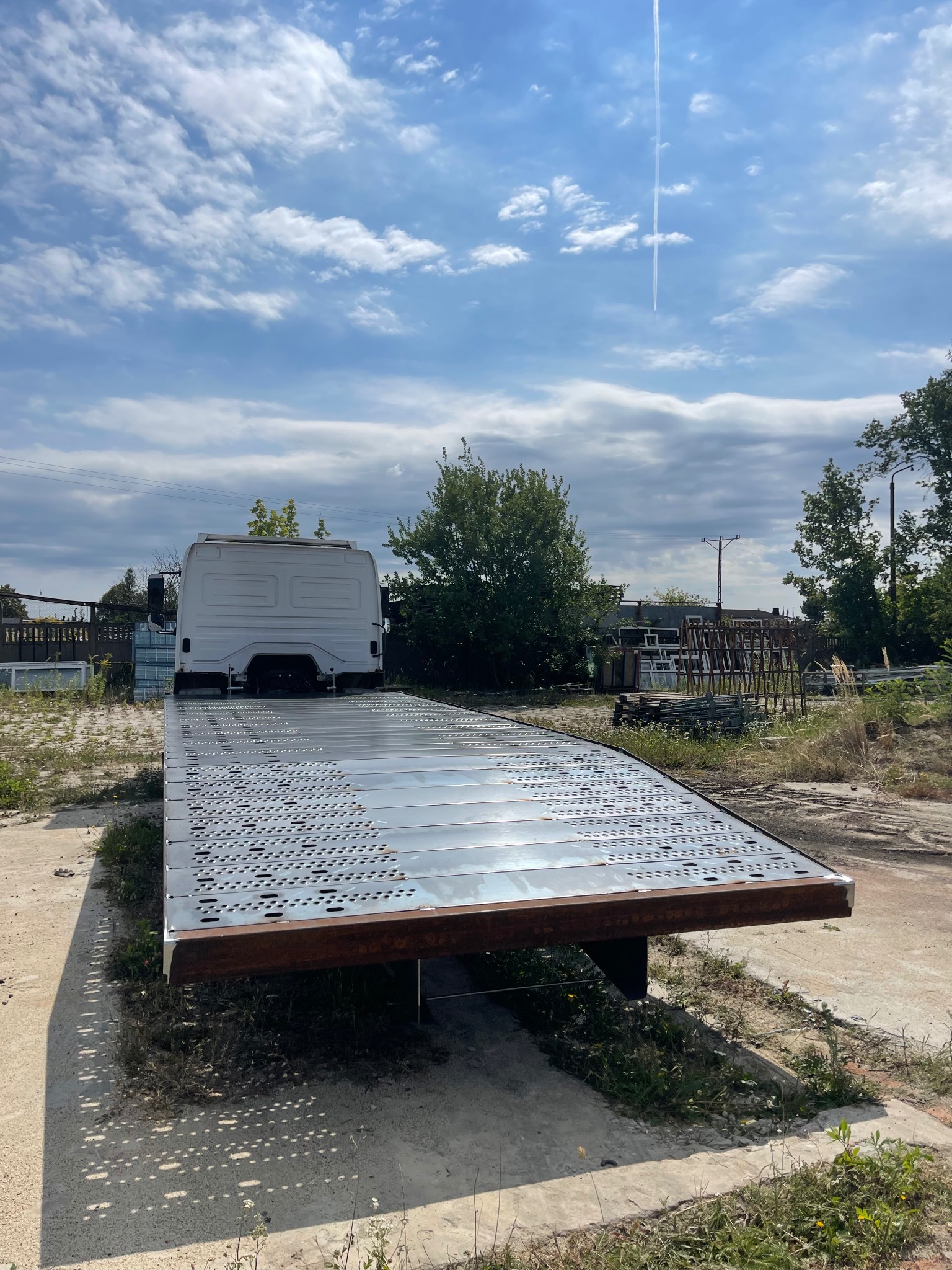 Platforma transportowa z perforowanej blachy, zamontowana na samochodzie dostawczym, na tle błękitnego nieba z chmurami i zieleni. Widok z tyłu pojazdu.
