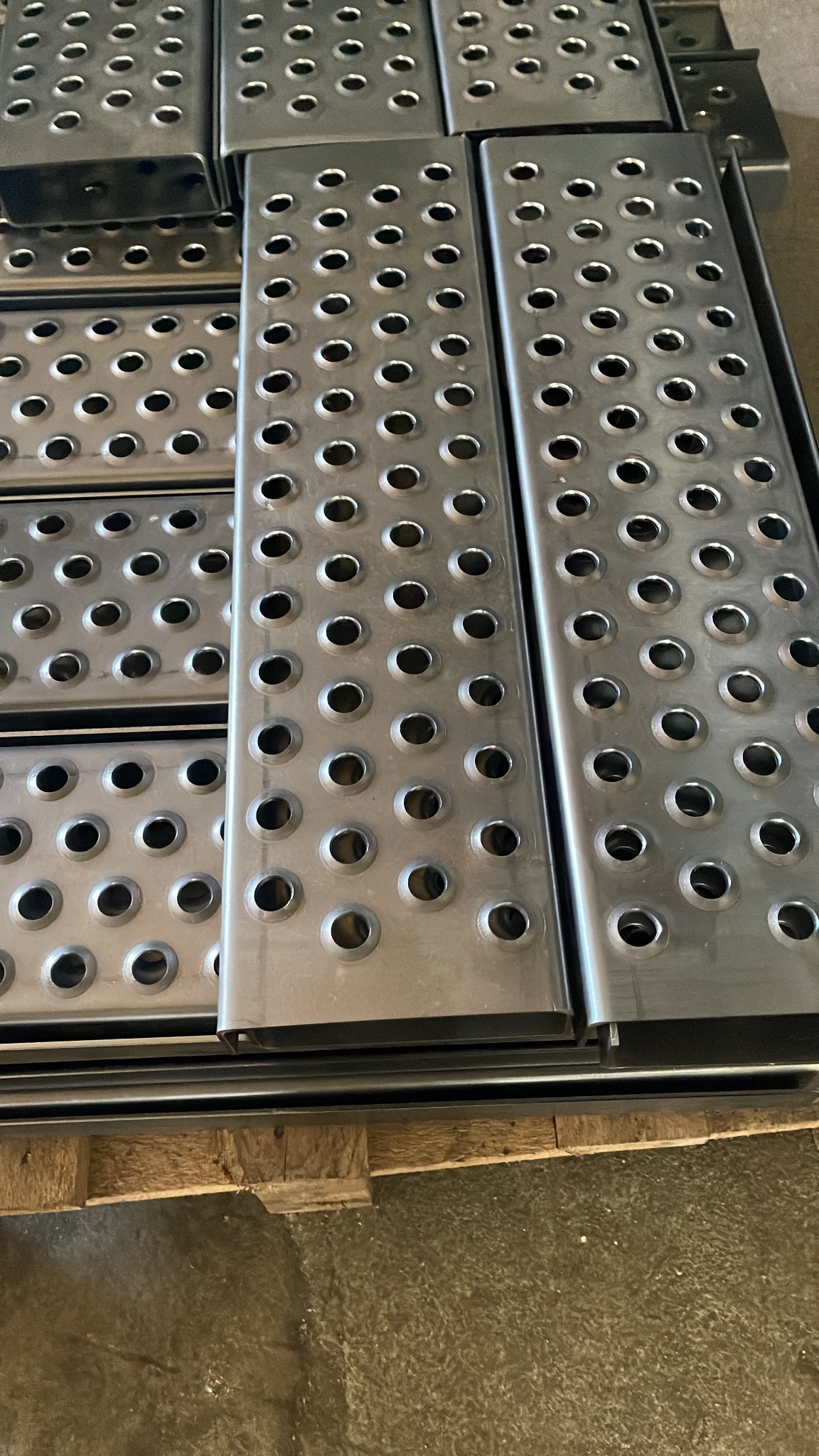 Metalowe stopnie z otworami, ułożone na palecie. Elementy konstrukcyjne ze stali, gotowe do montażu. Widoczne detale wykończenia i perforacja.