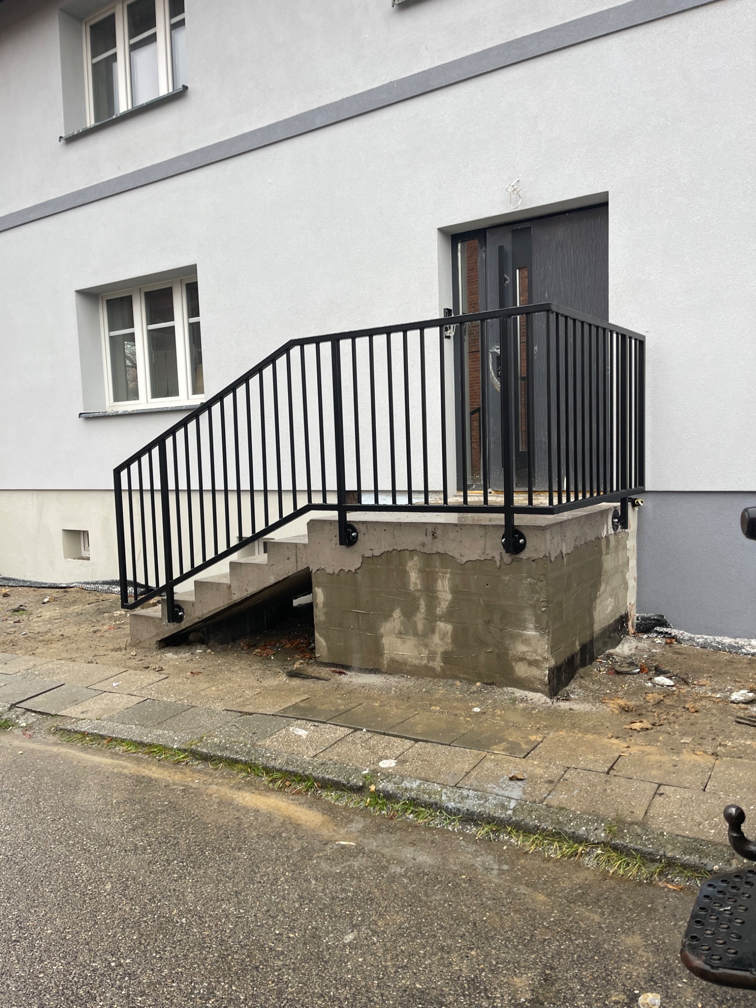 Czarne, metalowe poręcze schodowe zamontowane przy wejściu do budynku z szarą elewacją. Widoczne betonowe schody i fragment chodnika. Ujęcie z dołu.