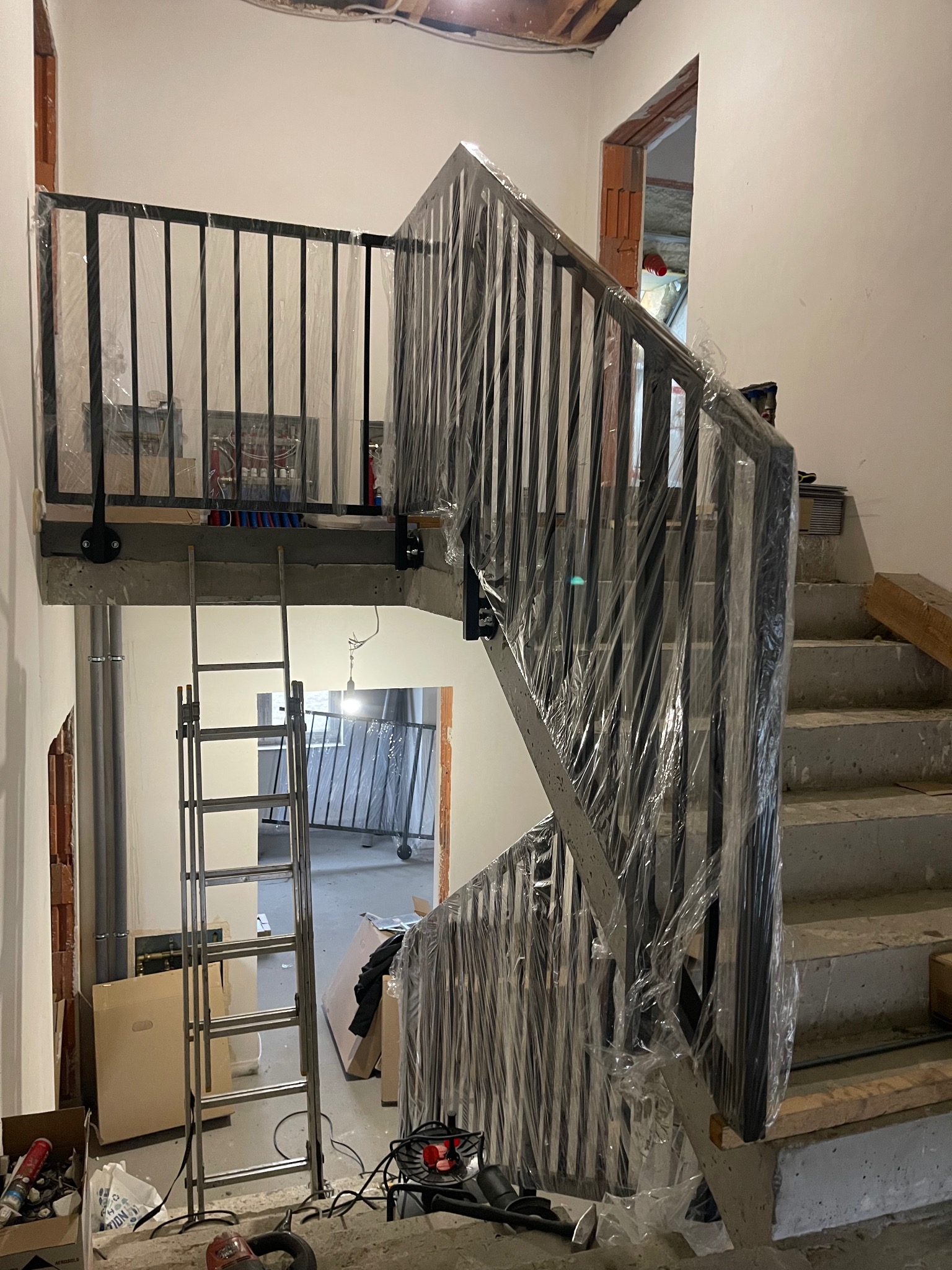 Czarne, metalowe schody z balustradą w folii ochronnej, w surowym wnętrzu. Widoczna drabina i elementy instalacji. Ujęcie z dołu, podkreślające konstrukcję schodów.