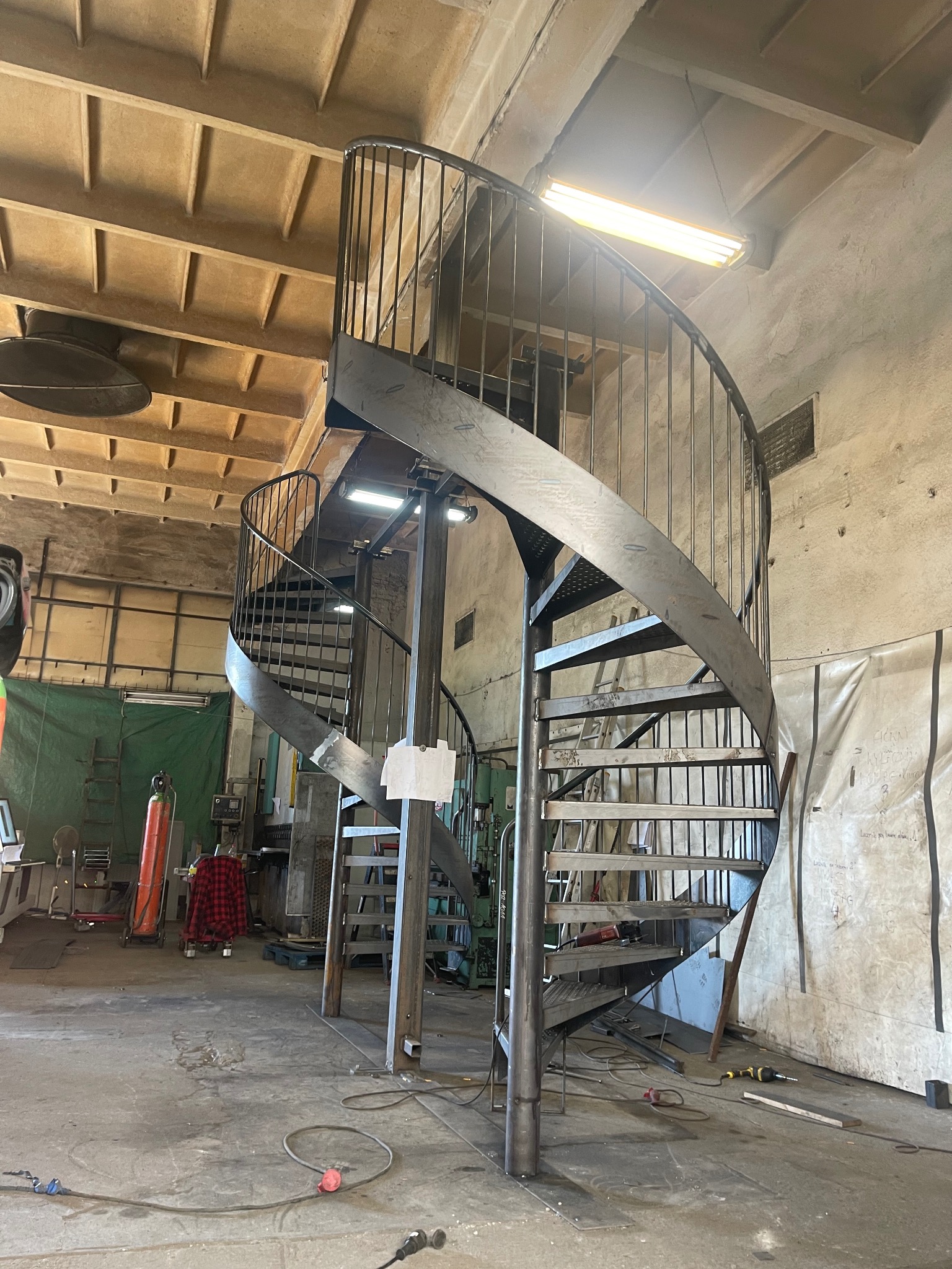 Metalowe schody spiralne w warsztacie, surowy metal, widoczne spawy, oświetlenie warsztatowe, narzędzia w tle, wysoki kąt ujęcia, ukazujący konstrukcję w trakcie montażu.