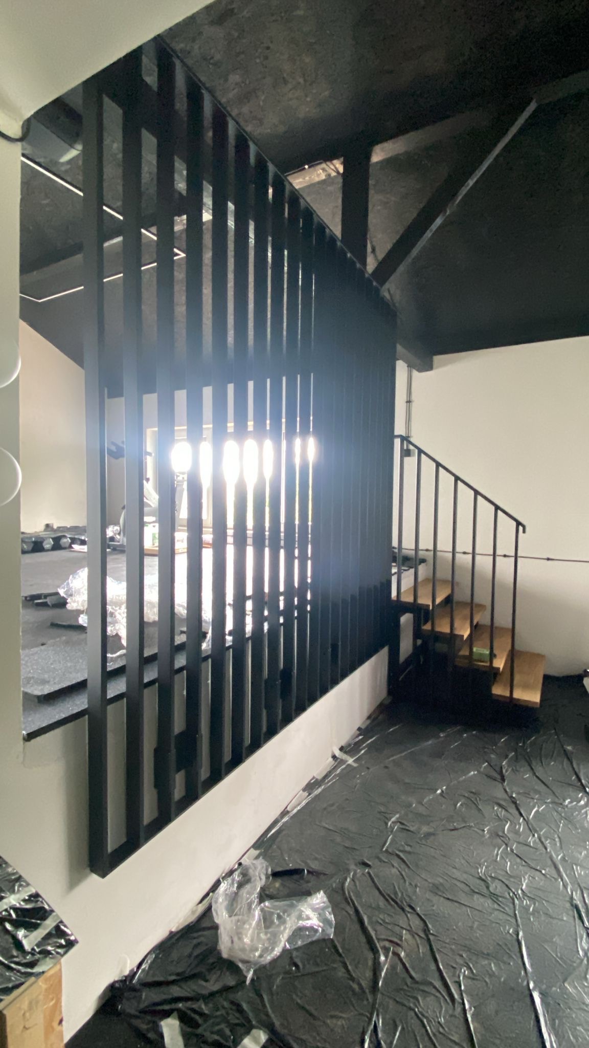 Nowoczesna, czarna balustrada z pionowych profili, oddzielająca przestrzeń. W tle schody z metalową poręczą. Podłoga zabezpieczona folią malarską.
