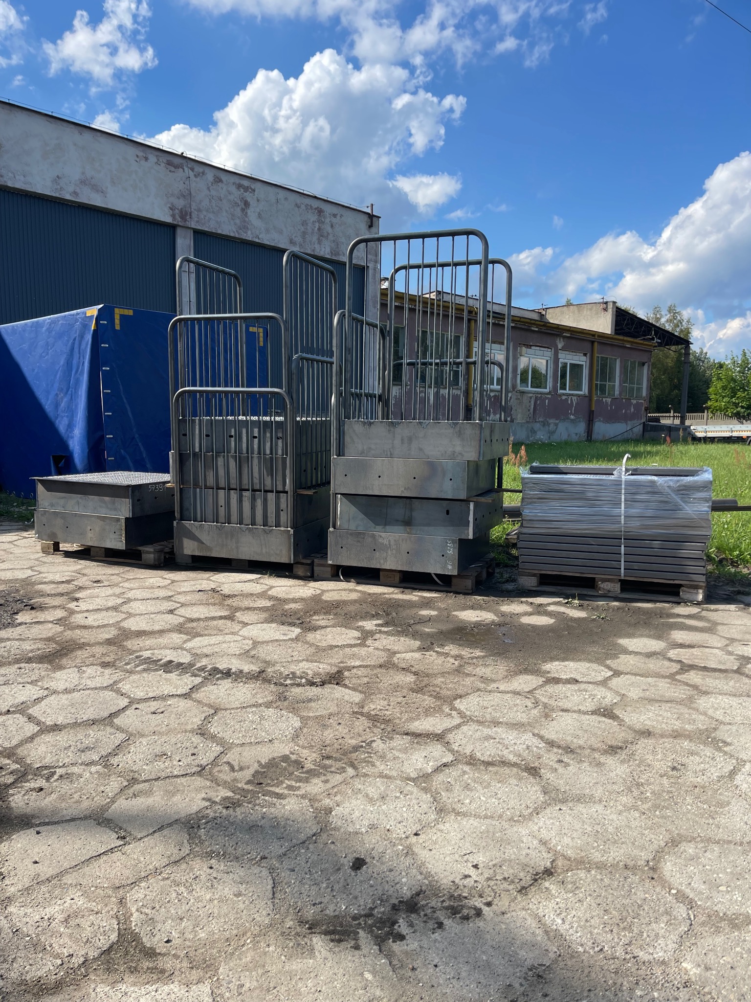 Stalowe konstrukcje schodkowe z poręczami, ułożone na paletach na zewnątrz budynku przemysłowego, w tle błękitne niebo z chmurami i budynek.