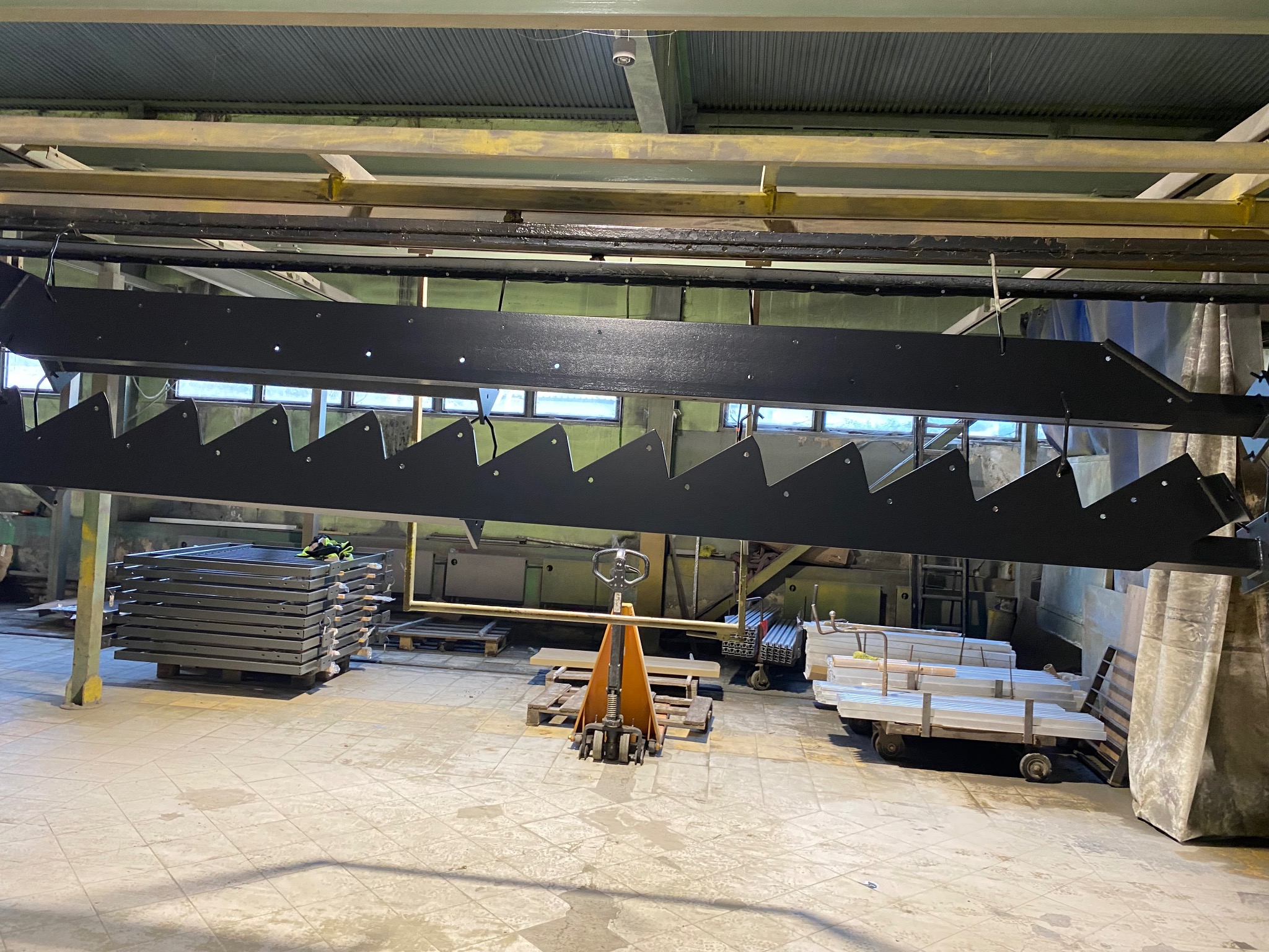 Czarne, metalowe stopnie schodowe zawieszone w hali produkcyjnej, czekające na dalszą obróbkę. W tle wózki z elementami i palety. Widoczne oświetlenie przemysłowe.