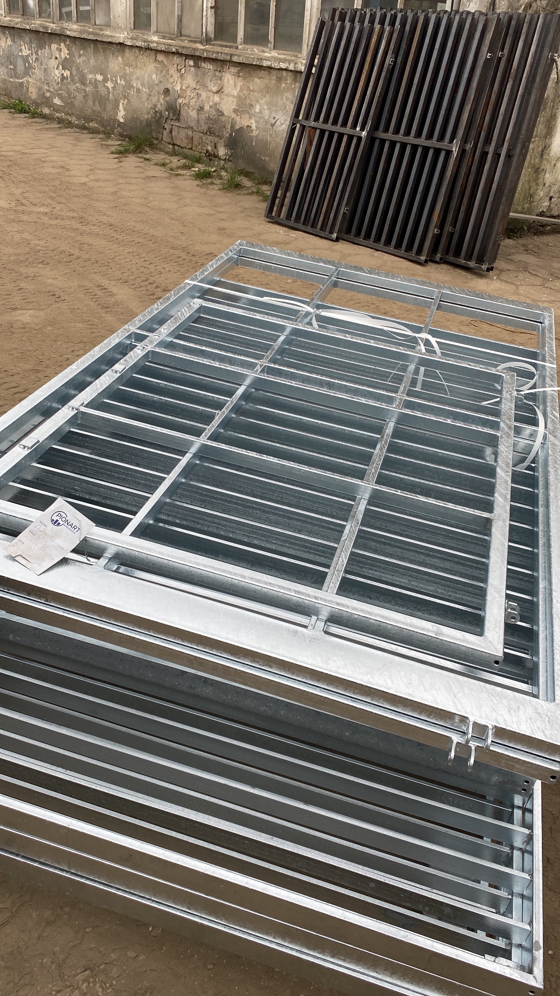 Stos ocynkowanych, spawanych krat pomostowych z etykietą producenta na ziemi. W tle widoczne kraty w kolorze czarnym i fragment budynku.