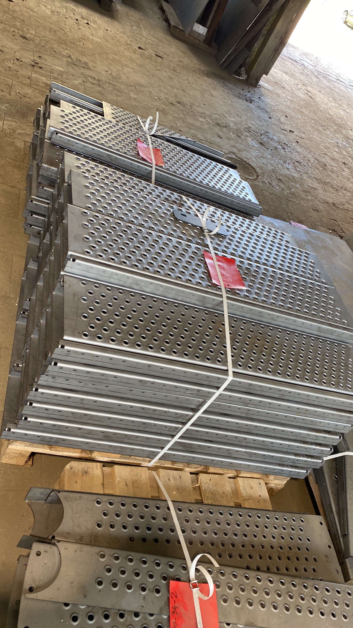 Stos metalowych, perforowanych stopni na palecie, zabezpieczonych sznurkiem i oznaczonych czerwoną etykietą. Widoczne otwory i krawędzie, surowy wygląd.