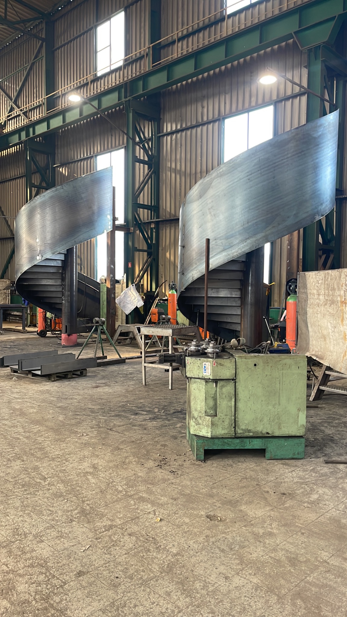 Wnętrze hali produkcyjnej z dwoma spiralnymi konstrukcjami schodów metalowych w trakcie budowy. Widoczne narzędzia i materiały, surowe wykończenie.