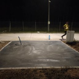 Kewin kuciarski - Świeżo wylana, betonowa powierzchnia otoczona ziemią, z widocznym narzędziem i butelką wody. W tle biegnąca osoba i oświetlenie uliczne w nocnej scenerii.