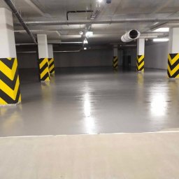 Kewin kuciarski - Gładka, szara posadzka w hali garażowej z kolumnami oznaczonymi czarno-żółtymi pasami ostrzegawczymi. Widoczne elementy wentylacji pod sufitem.