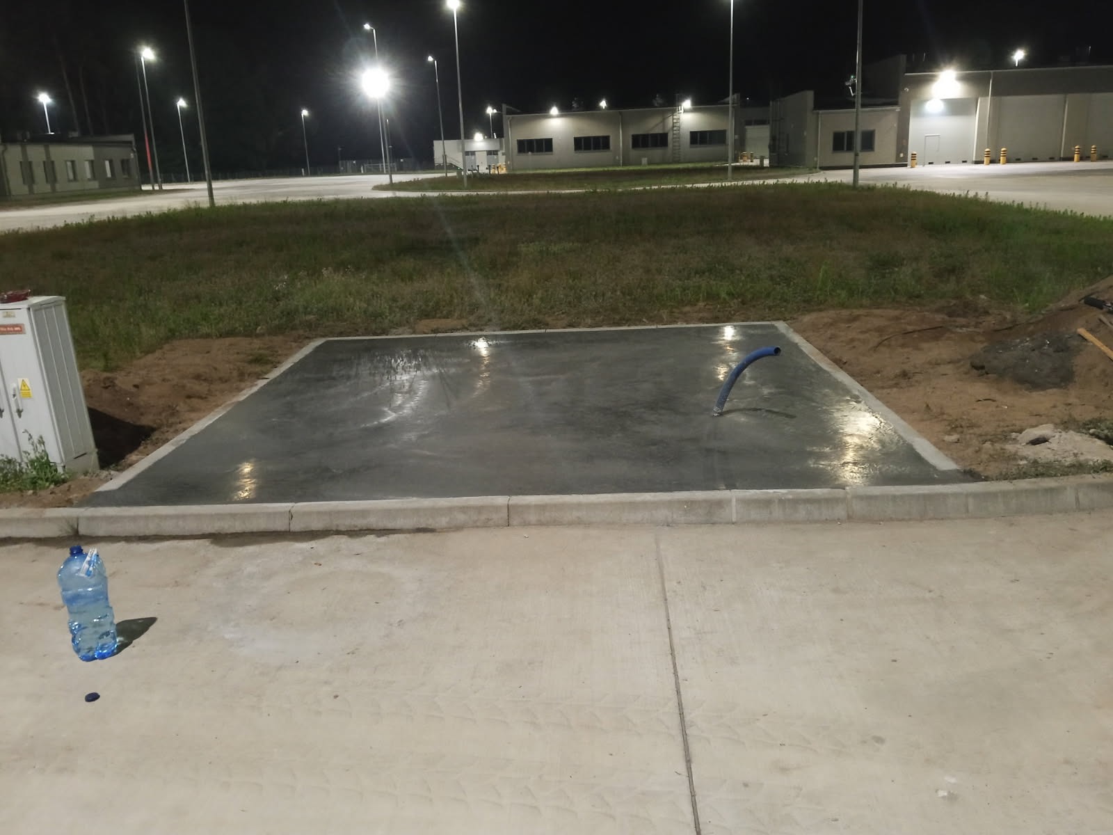 Wylana, mokra, betonowa posadzka przemysłowa na zewnątrz, otoczona kostką. W tle budynki przemysłowe i oświetlenie nocne. Widoczny niebieski wąż i butelka z wodą.