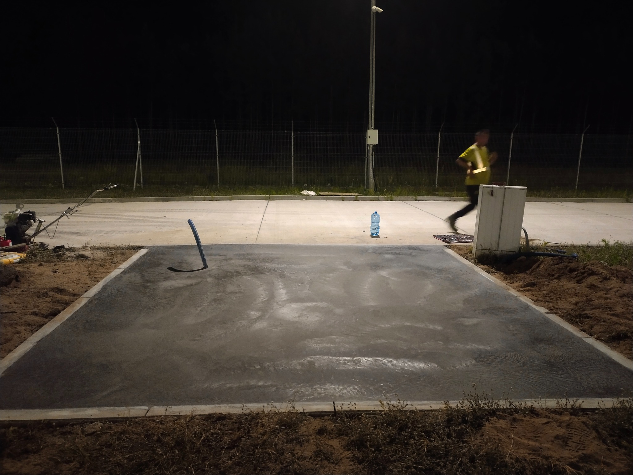 Świeżo wylana, betonowa powierzchnia otoczona ziemią, z widocznym narzędziem i butelką wody. W tle biegnąca osoba i oświetlenie uliczne w nocnej scenerii.