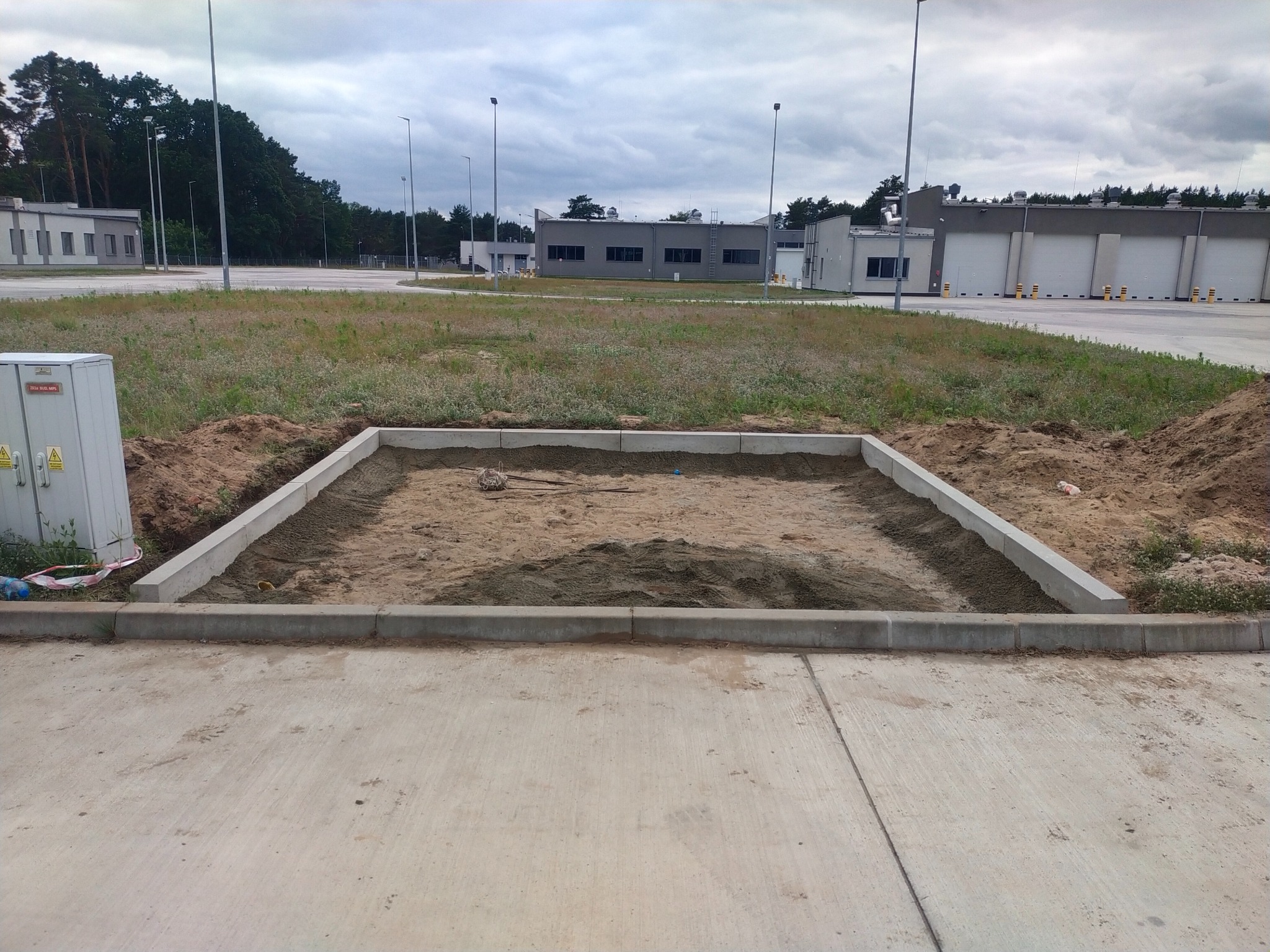 Przygotowanie podłoża pod posadzkę przemysłową z obrzeżami betonowymi na zewnątrz budynku. Widoczne budynki w tle i pochmurne niebo.