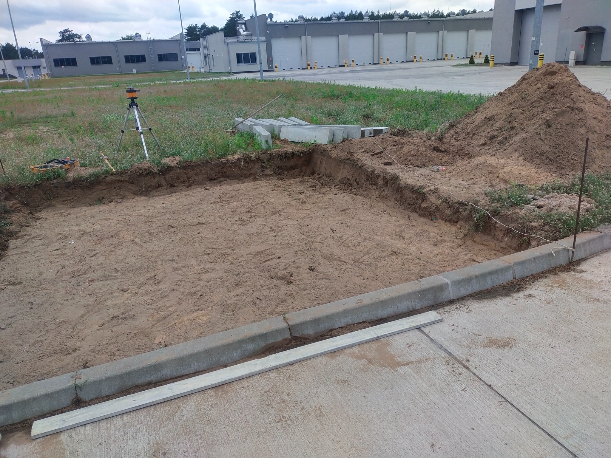 Przygotowanie terenu pod posadzkę: wykop z piaskiem, obrzeża z betonowych krawężników, niwelator laserowy na statywie, sterta ziemi w tle.