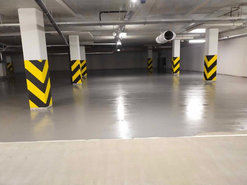 Gładka, szara posadzka w hali garażowej z kolumnami oznaczonymi czarno-żółtymi pasami ostrzegawczymi. Widoczne elementy wentylacji pod sufitem.