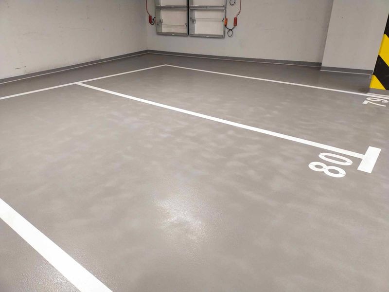 Szara posadzka przemysłowa z białymi liniami i numerami miejsc parkingowych w garażu podziemnym. Widoczne zabezpieczenia słupa.