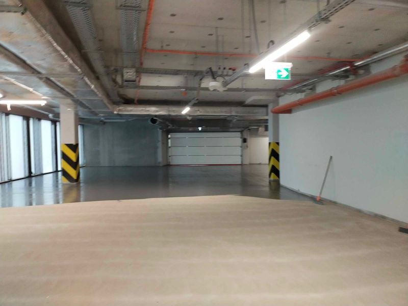 Gładka, jasna posadzka w hali garażowej z kolumnami oznaczonymi pasami ostrzegawczymi. Widoczny betonowy sufit z instalacjami i brama garażowa.