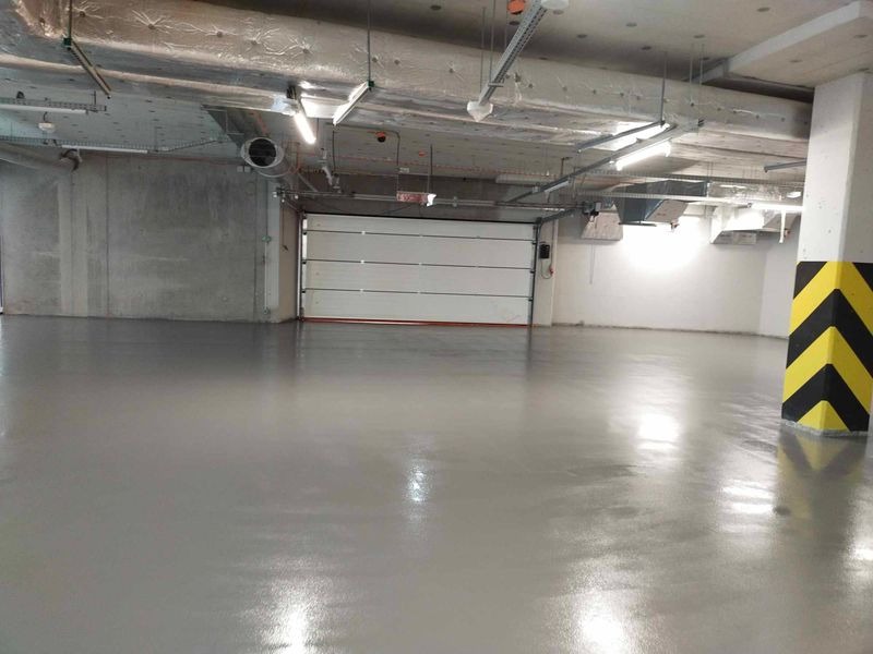 Gładka, szara powierzchnia w hali garażowej w Zielonej Górze, z widocznymi elementami instalacji pod sufitem i kolumną w żółto-czarne pasy.