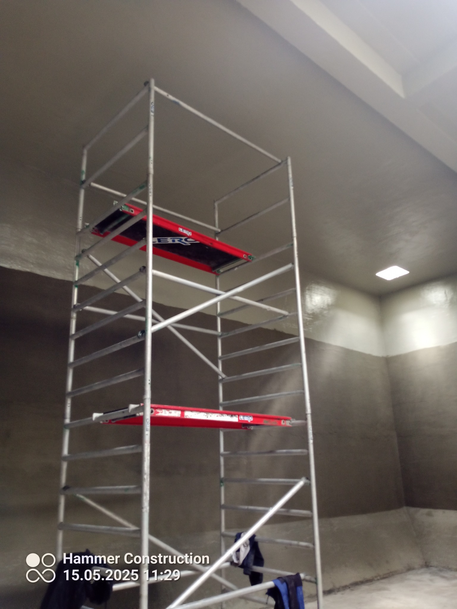 Wnętrze w trakcie prac wykończeniowych: metalowe rusztowanie na tle świeżo pomalowanej ściany w odcieniach szarości, widoczne niedoskonałości i różnice w kolorze.