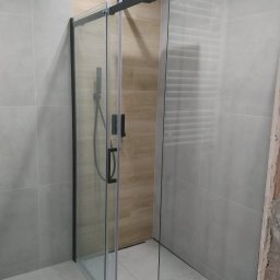 EXECUTIVE COOPERATION COMPANY E.C.C_D&S - Nowoczesna kabina prysznicowa z przesuwnymi drzwiami, czarne profile, szklane ściany, jasne płytki w łazience. Widoczny fragment ściany w trakcie wykończenia.