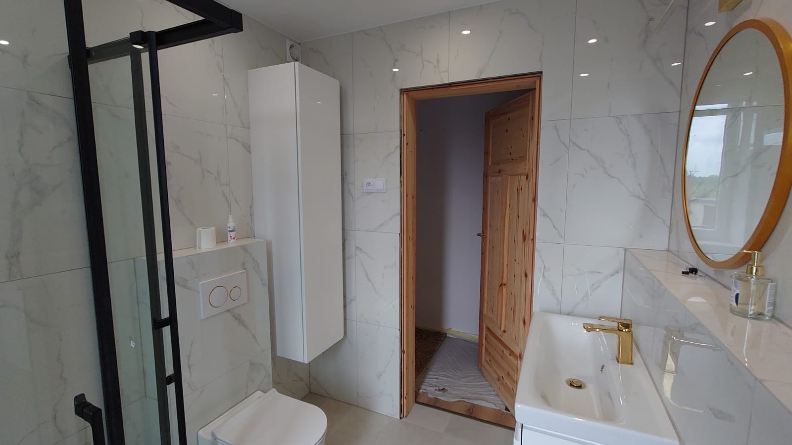 Wykończona łazienka z kabiną prysznicową typu walk-in, białą armaturą i płytkami imitującymi marmur. Złota bateria umywalkowa i okrągłe lustro dodają elegancji.