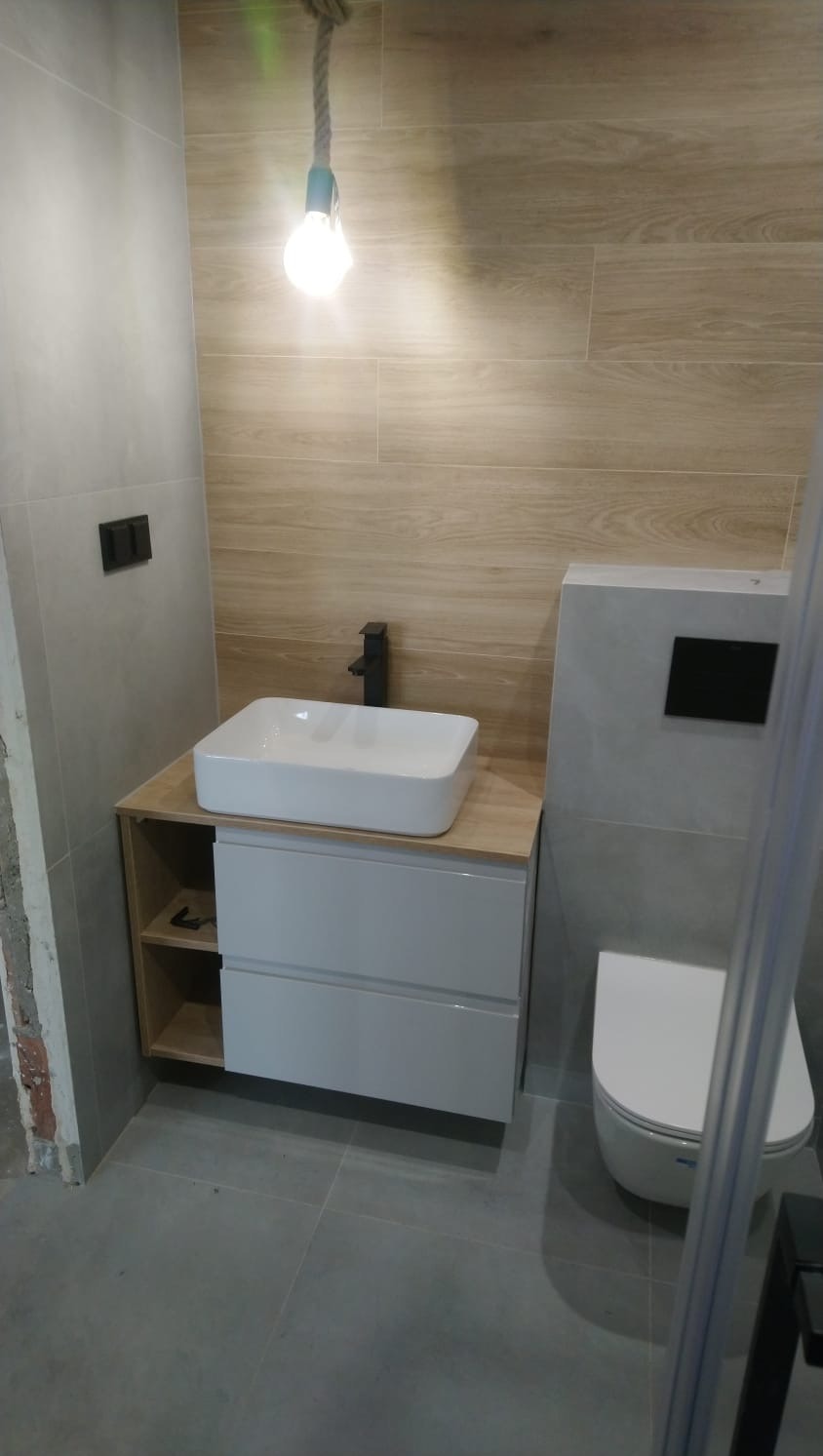 Nowoczesna łazienka po remoncie: biała umywalka nablatowa z czarną armaturą, drewnopodobne płytki ścienne, szara podłoga i wisząca toaleta. Minimalistyczny design.