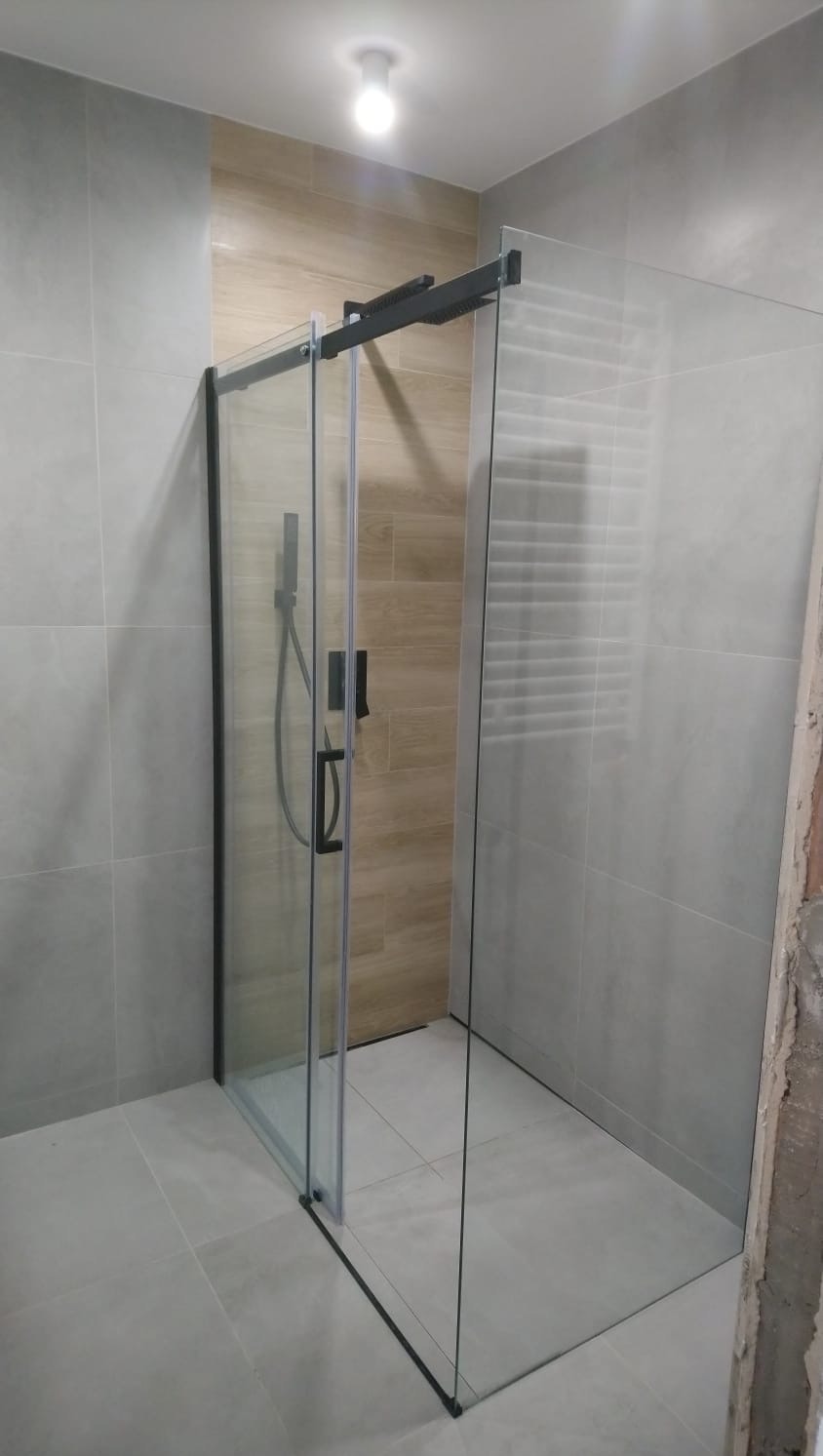 Nowoczesna kabina prysznicowa z przesuwnymi drzwiami, czarne profile, szklane ściany, jasne płytki w łazience. Widoczny fragment ściany w trakcie wykończenia.