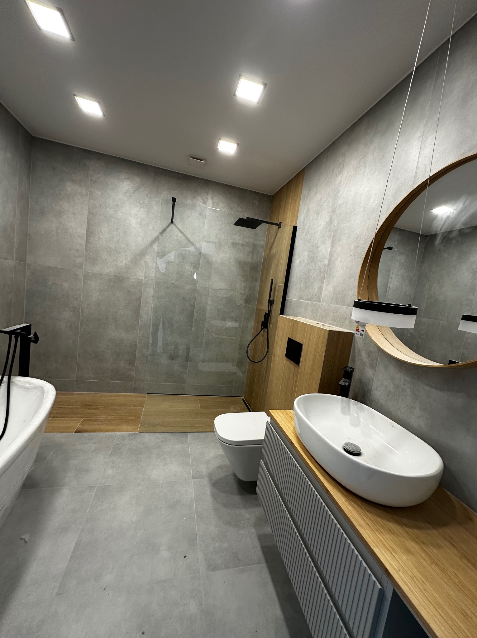 Nowoczesna łazienka z szarymi płytkami, drewnianymi akcentami i okrągłym lustrem. Kabina prysznicowa typu walk-in z czarną armaturą. Umywalka nablatowa na drewnianym blacie.