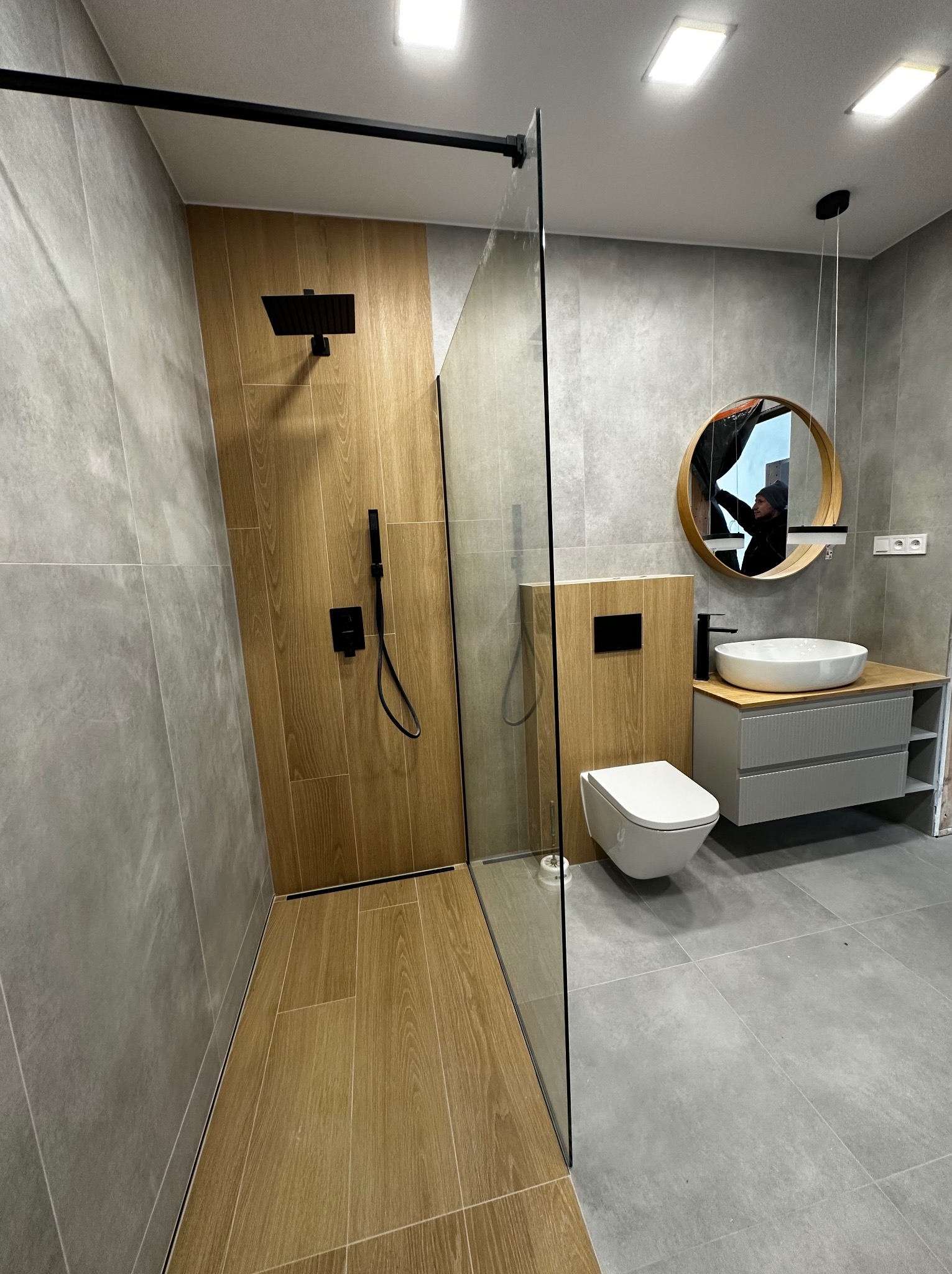 Nowoczesna łazienka z kabiną typu walk-in, drewnopodobnymi płytkami w strefie prysznica i szarą ceramiką na ścianach. Okrągłe lustro i minimalistyczna umywalka.