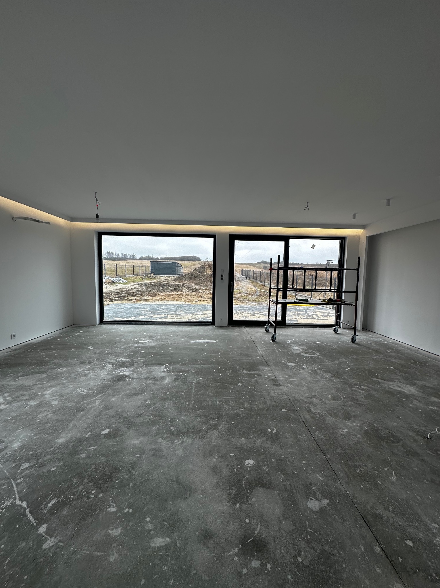 Surowe wnętrze z widokiem na zewnątrz przez duże okna. Na podłodze widoczne ślady po pracach. W tle rusztowanie malarskie. Minimalistyczny design z oświetleniem LED.