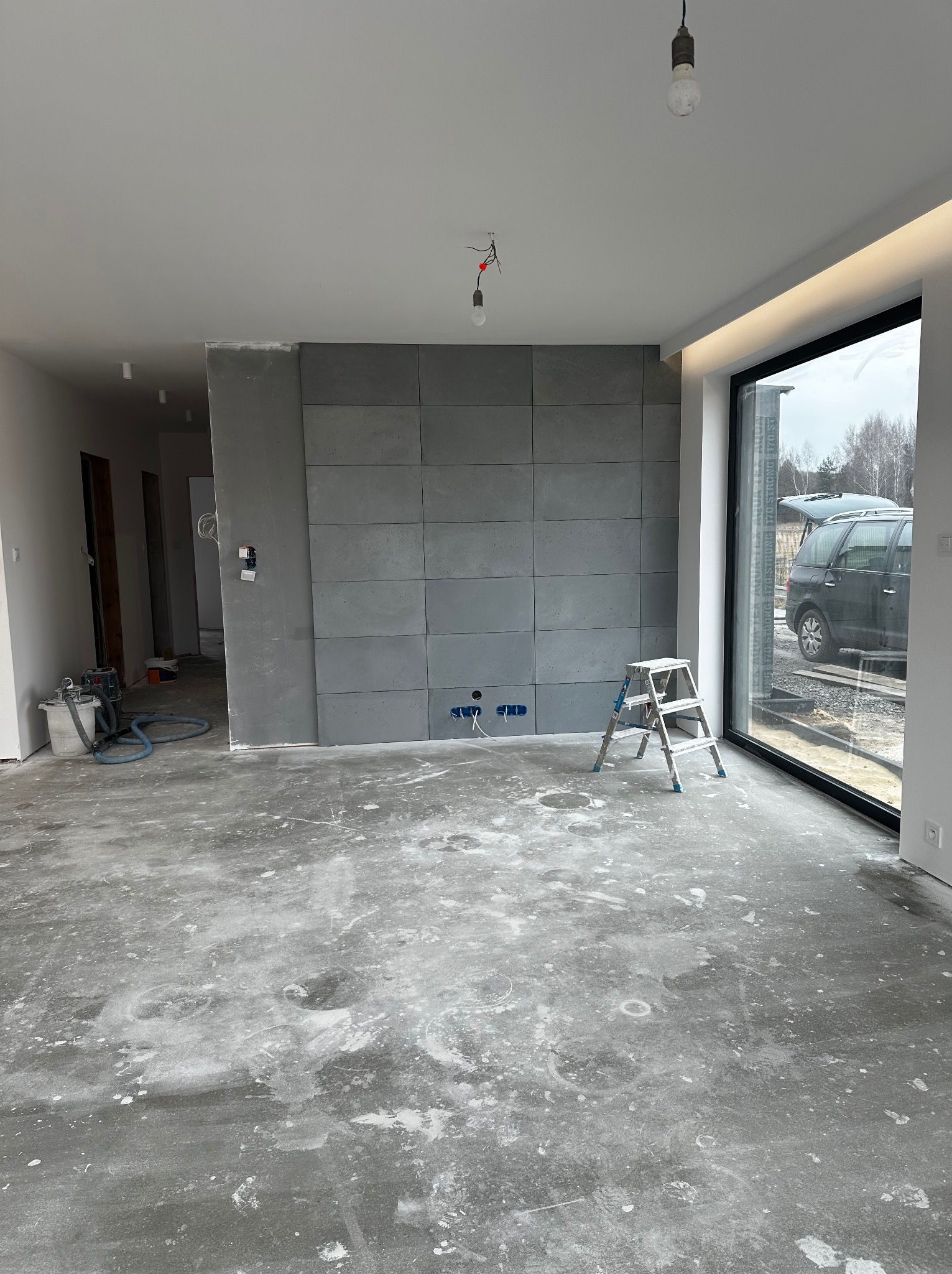 Wnętrze w trakcie prac: ściana z betonowych płyt, aluminiowa drabina, surowa podłoga z resztkami zaprawy, duże okno z widokiem na zewnątrz. Widoczne instalacje elektryczne.